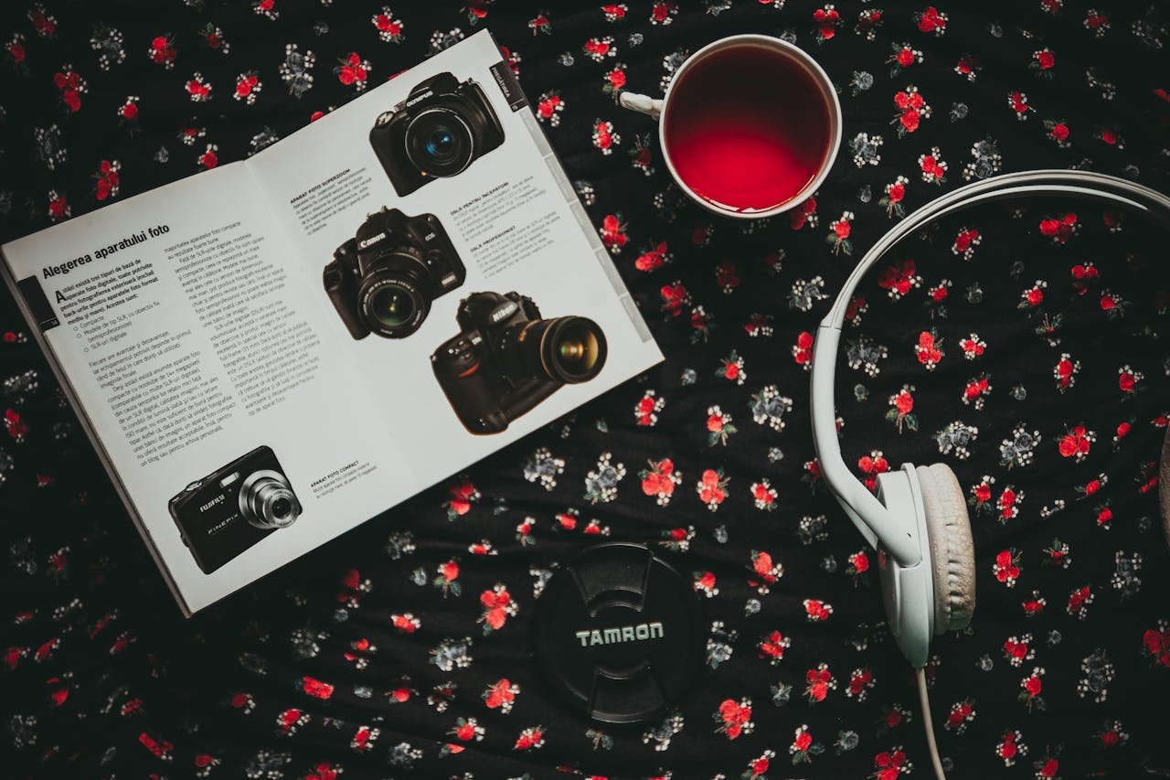 Flat lay composition of camera gear, headphones, magazine, and tea cup on floral background.
